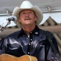 Ein Country-Sänger mit Cowboyhut spielt eine Akustikgitarre und singt vor einem Mikrofon auf einer Bühne.