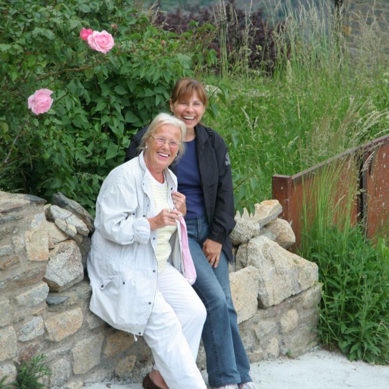 Zwei lächelnde Frauen sitzen draußen an einer Steinmauer, umgeben von üppigem Grün und blühenden Rosen.