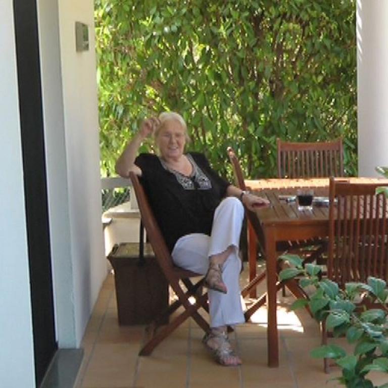 Eine lächelnde ältere Frau sitzt draußen auf einer Terrasse vor einem Hintergrund aus üppigem Grün.