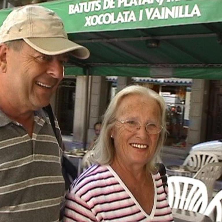 Ein lächelnder Mann und eine ältere Frau stehen vor einem Marktstand.