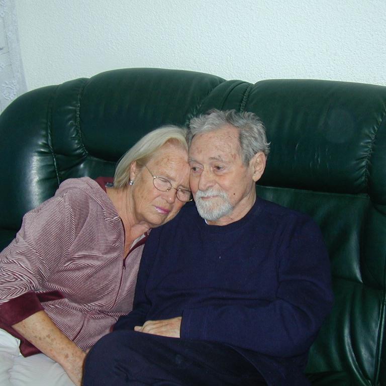 Ein älteres Paar sitzt auf einem dunklen Ledersofa und blickt freundlich in die Kamera.