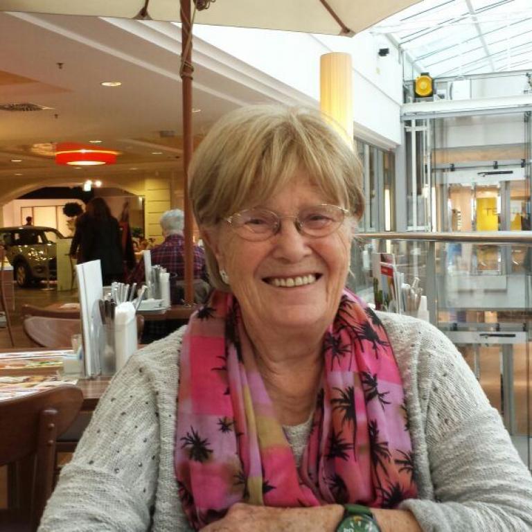 Eine lächelnde Frau steht in einem hell beleuchteten, modernen Innenraum, der wie ein Einkaufszentrum wirkt.