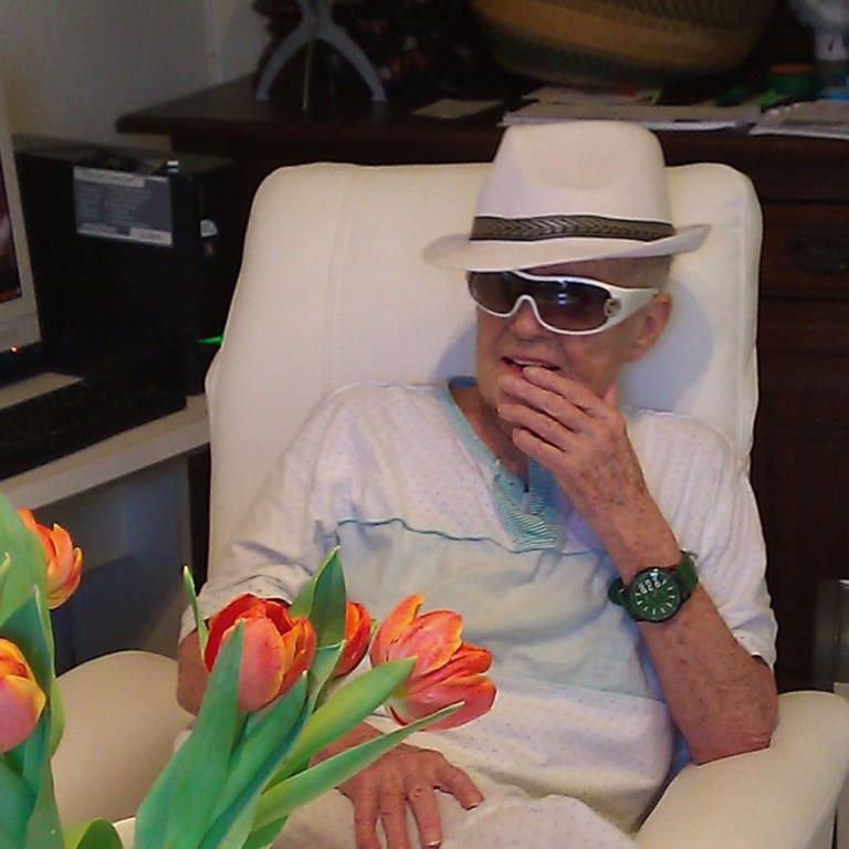 Ein Mann in Hut und Sonnenbrille sitzt in einem Bürostuhl und blickt nachdenklich, mit einem Blumenstrauß im Vordergrund.