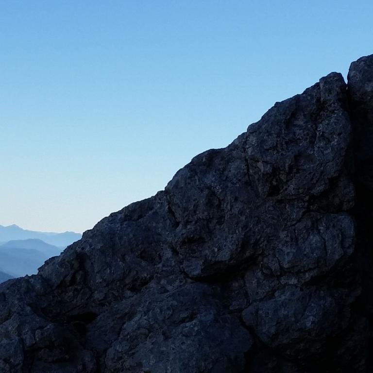 Ein dunkler Felsvorsprung im Vordergrund rahmt einen weiten Blick auf eine Berglandschaft mit blauem Himmel ein.