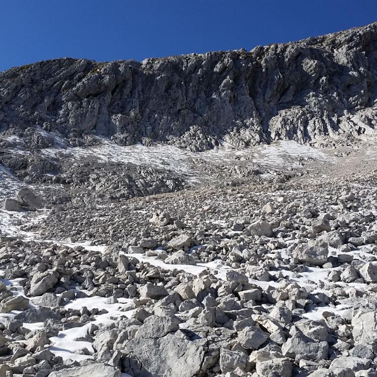 Eine weite Berglandschaft mit Schutthalde, Schnee und einem steilen Felsabhang unter blauem Himmel.