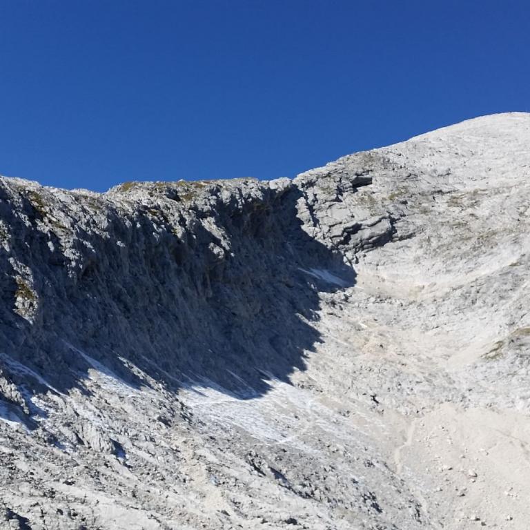 Steil abfallende Felswand und Geröllfeld in einer Hochgebirgslandschaft unter blauem Himmel.