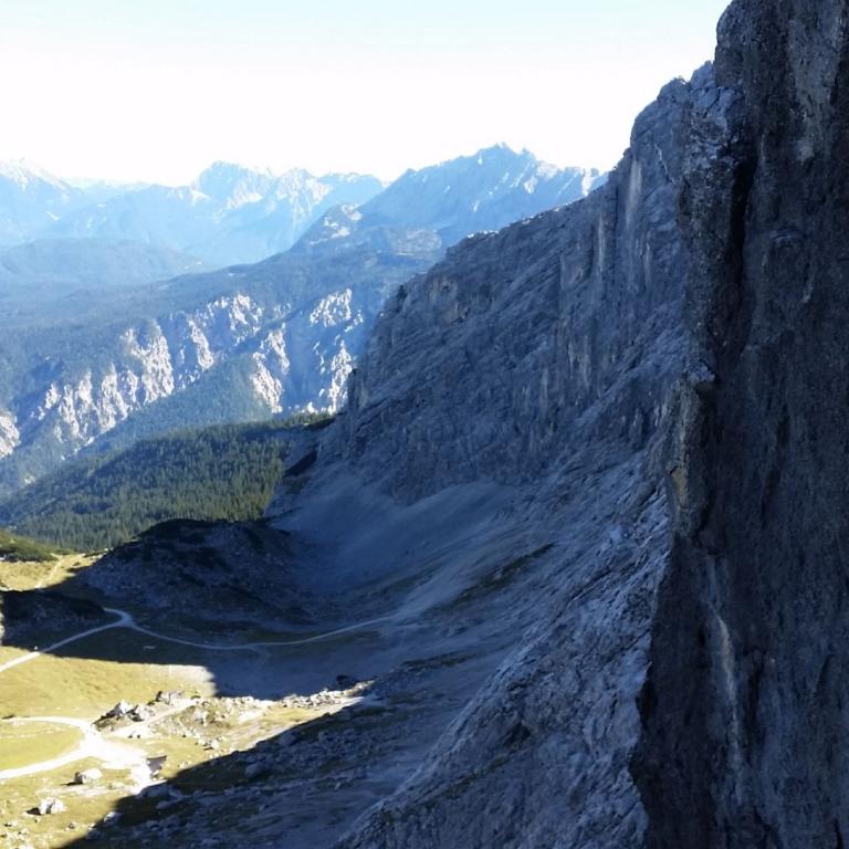 Eine hohe, dunkle Felswand stürzt über ein weitläufiges Tal mit Blick auf entfernte, bergige Landschaften.