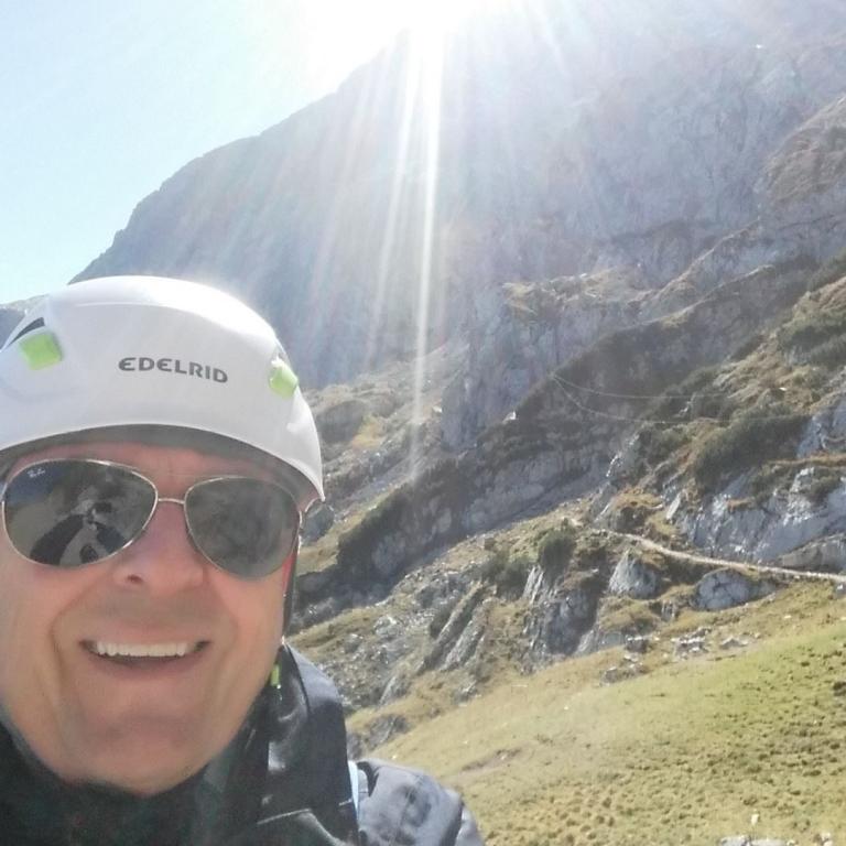 Ein Mann macht ein Selfie vor einer bergigen Landschaft bei hellem Sonnenschein.