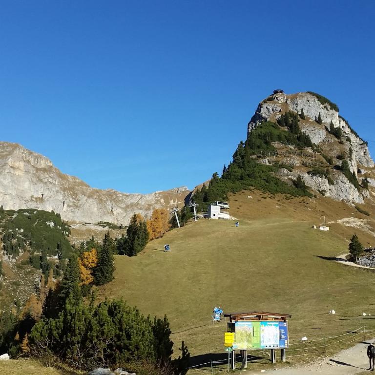 Eine weite Berglandschaft mit einem markanten Felsgipfel in der Mitte und einer grünen Almwiese unter blauem Himmel.