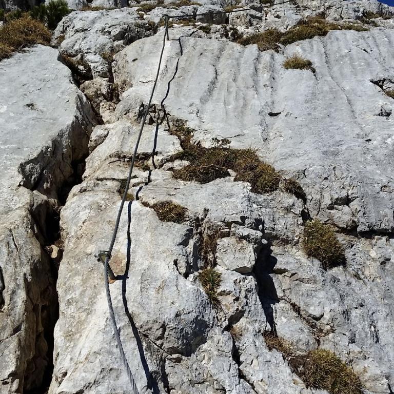 Ein Kletterweg mit Seilen führt an einer steilen Felswand hinauf zu einer felsigen Bergkuppe.