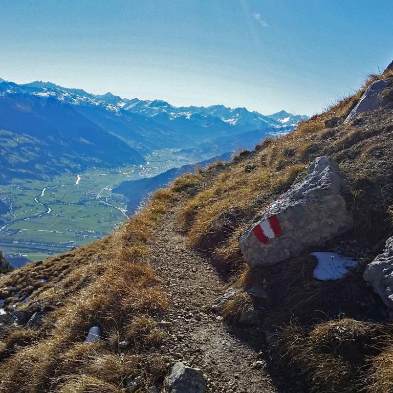 Ein Wanderweg führt über einen Berghang mit einem markanten Wegweiser und bietet einen weiten Blick auf ein Tal und dahinterliegende Bergketten.
