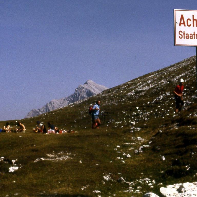 Eine Gruppe von Menschen auf einer bergigen Wiese unter einem blauen Himmel, mit einem Warnschild für Staatsgrenze.