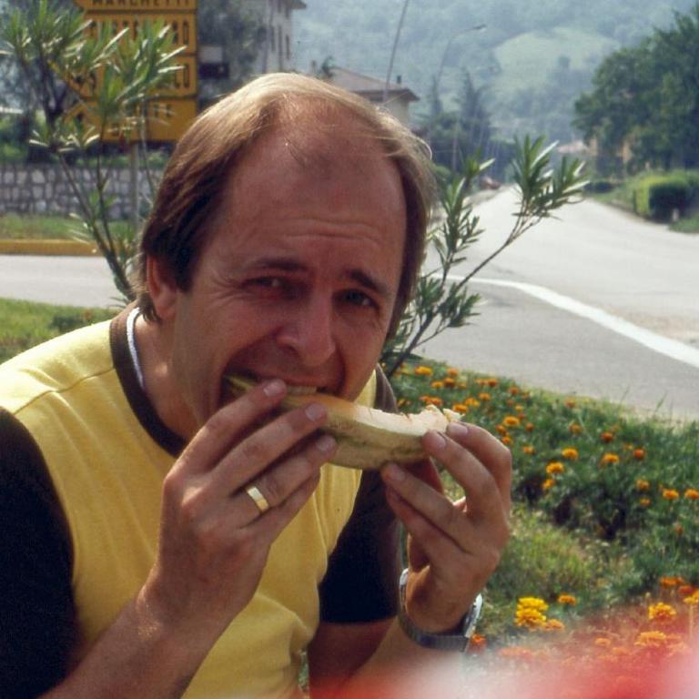 Ein Mann sitzt im Freien und isst ein Stück Nahrung, mit einer Straße und Grün im Hintergrund.