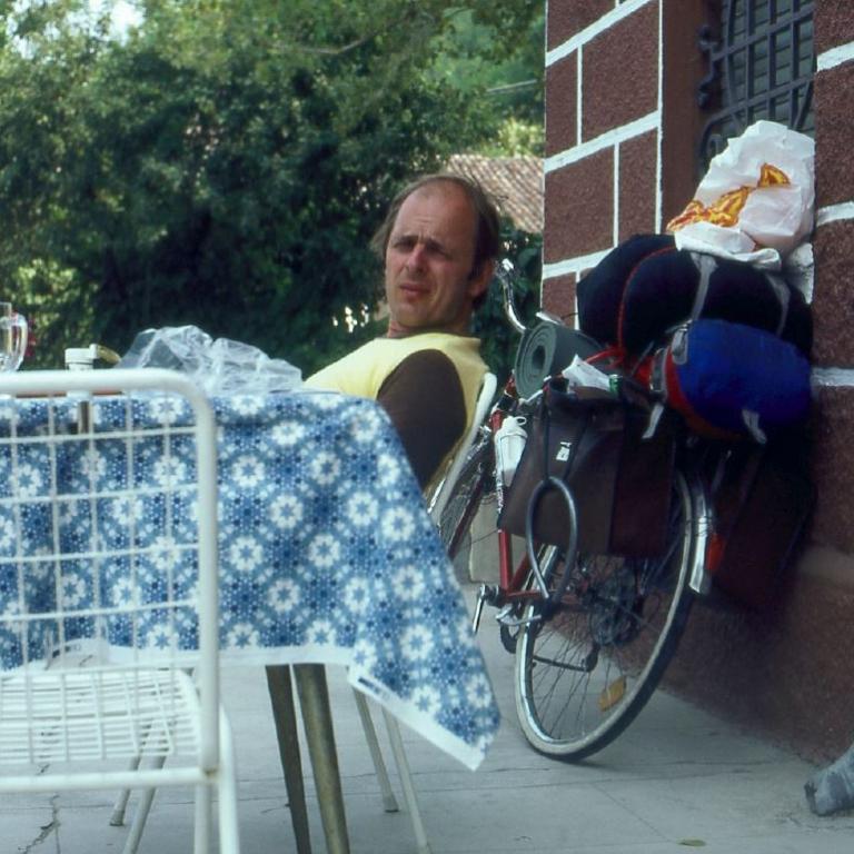 Ein Mann sitzt an einem Picknicktisch im Freien, neben dem steht ein Fahrrad, das mit Taschen beladen ist.