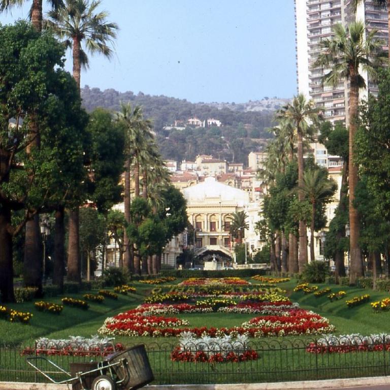 Ein weitläufiger städtischer Park mit vielen Palmen, blühenden Blumenbeeten und historischen Gebäuden im Hintergrund.