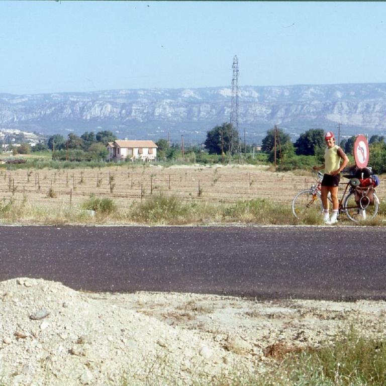 Mann mit Fahrrad an einer Straße vor einer weiten Landschaft mit Feldern und Bergen.