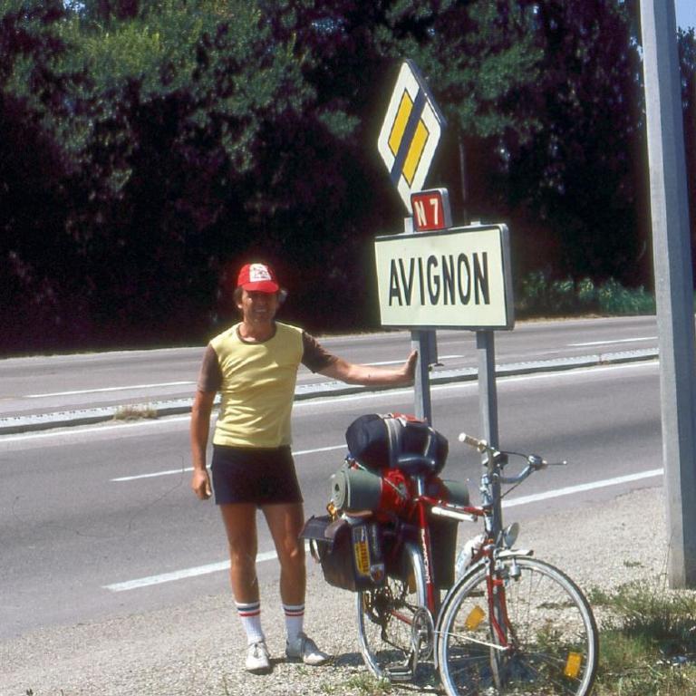 Ein Mann steht neben einem Fahrrad vor einem Straßenschild, das Richtung Avignon weist, an einer Hauptverkehrsstraße.
