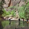 Ein ruhiger Fluss fließt durch einen Canyon, der von hohen Felswänden und üppigem Wald umgeben ist. Einige Menschen stehen am bewaldeten Ufer und geni