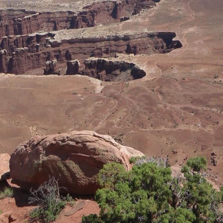 Panorama einer kargen Wüstenlandschaft mit tiefen Canyons, geschichteten roten Felswänden und großen Felsbrocken im Vordergrund.